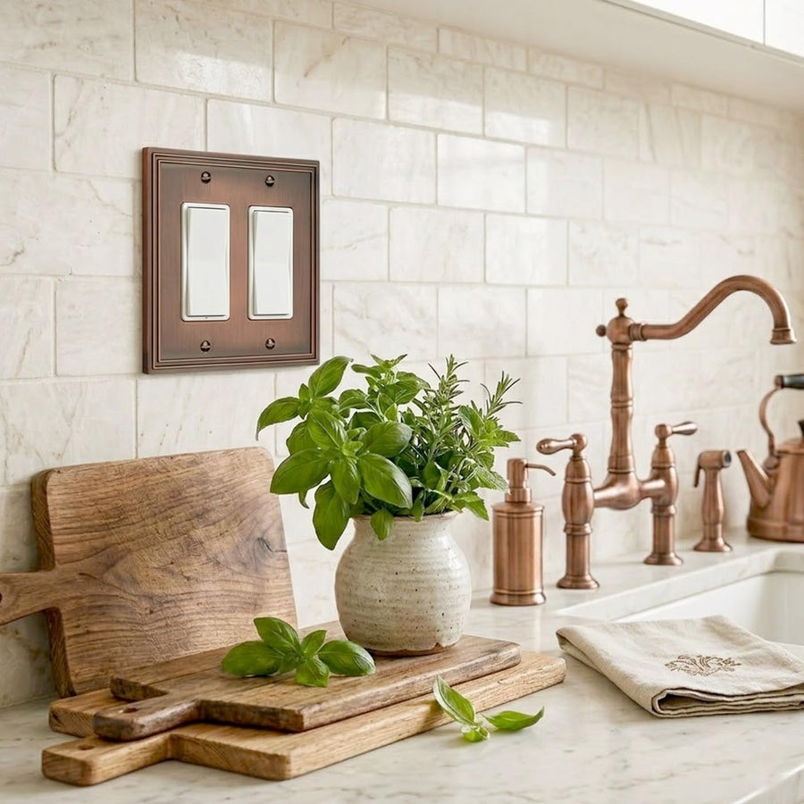 Imperial 2-gang rocker switch plate in Vintage Copper on a white tile backsplash beside a copper faucet and cutting boards