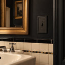 Black Pearl toggle switch plate with beaded border and a black toggle on a charcoal bathroom wall above cream tile with a black and gold border.