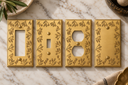 Four Floral Brushed Gold switch plates on marble: a rocker plate, a toggle plate, a duplex outlet cover, and a blank plate.