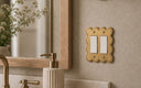 A Scallop 2G Rocker switch plate in Brushed Gold, angled on a soft beige bathroom wall beside an oak mirror and a brass soap pump