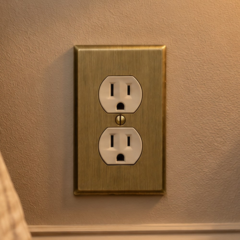 Signature Aged Gold duplex outlet cover above a baseboard beside a wood nightstand in a bedroom.