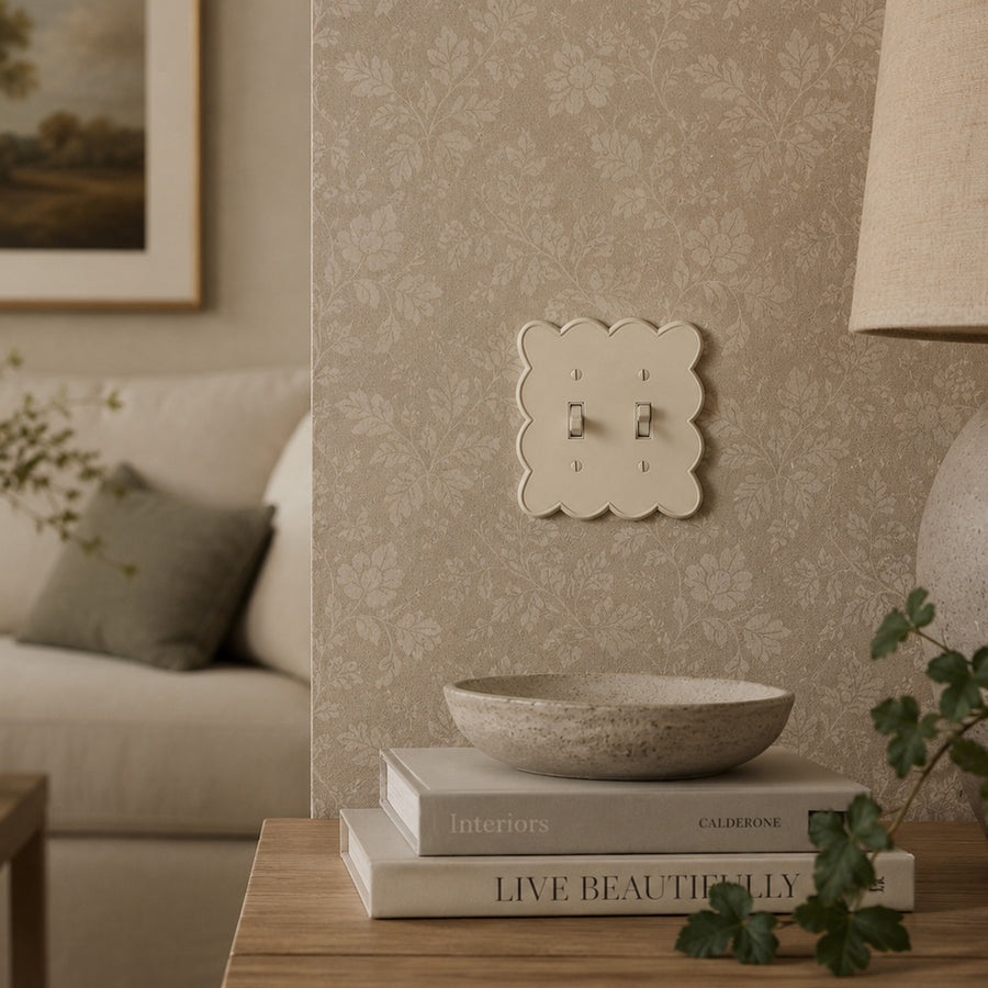 Scallop 2-gang toggle switch plate in Warm Ivory on a floral wallpaper wall above books and a stone bowl